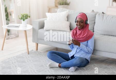 Restez à la maison. Femme musulmane noire souriante se détendant avec un smartphone et un casque Banque D'Images