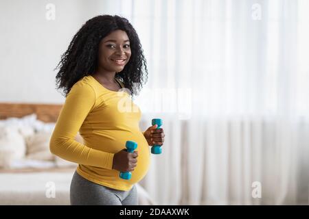 Bonne femme enceinte noire levant des haltères, s'exerçant à la maison Banque D'Images