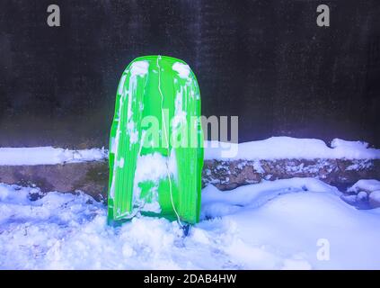 Traîneau en plastique vert sur de la neige fraîche près de la maison rurale mur à la campagne Banque D'Images