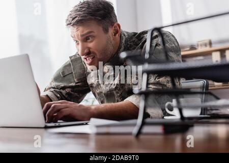 un militaire irrité tape sur le clavier de l'ordinateur portable près du plateau porte-documents sur un premier plan flou Banque D'Images