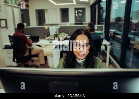 Femme d'affaires souriante de course mixte portant des lunettes de travail tard dans la journée saisie du rapport de finition au bureau sur l'ordinateur de bureau Banque D'Images