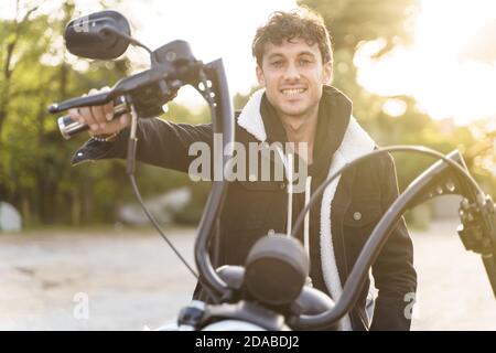 Portrait d'un jeune homme dans une veste souriante et assis sur une moto avec ses mains sur le guidon et face à la caméra avec les rayons du soleil shi Banque D'Images