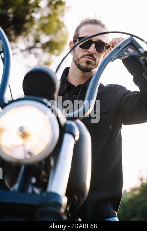 Photo verticale d'un jeune homme assis sur une moto tout en prenant ses lunettes de soleil entourées d'arbres Banque D'Images