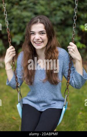 Jolie jeune fille assise sur balançoire souriante avec de la verdure dedans arrière-plan Banque D'Images