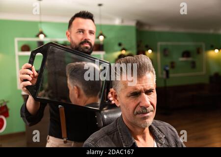 barber barbu montre la coupe de cheveux et la coiffure à son client avec un bouc avec un miroir dans le barbershop, les deux caucasiens. Banque D'Images