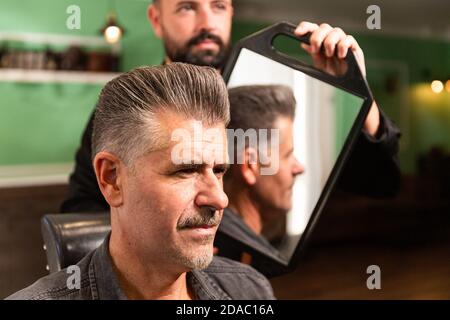 barber barbu montre la coupe de cheveux et la coiffure à son client avec un bouc avec un miroir dans le barbershop, les deux caucasiens. Banque D'Images