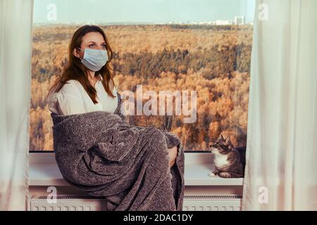 Une jeune femme s'assoit sur une bordure de fenêtre dans un masque médical en quarantaine. Concept de quarantaine du coronavirus et d'isolement à domicile Banque D'Images