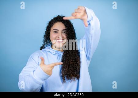 Jeune belle femme portant un sweat-shirt décontracté sur fond bleu isolé sourire en faisant le cadre avec les mains et les doigts avec le visage heureux Banque D'Images