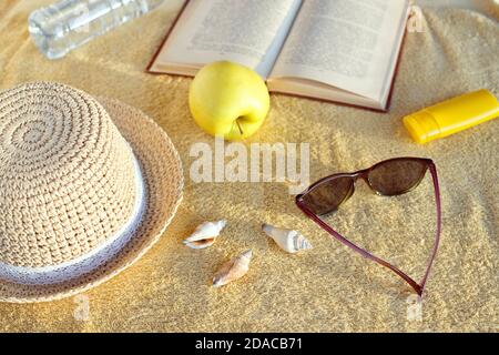 Chapeau de paille, coquillages, crème solaire, lunettes de soleil, pomme, bouteille d'eau et livre ouvert sur serviette de plage beige. Lumière douce du soleil le soir. Banque D'Images