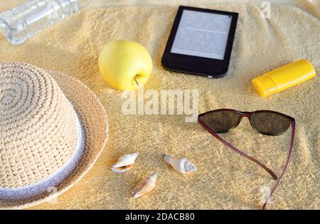 Chapeau de paille, coquillages, crème solaire, lunettes de soleil, pomme, bouteille d'eau et livre électronique sur serviette de plage beige. Lumière douce du soleil le soir. Banque D'Images