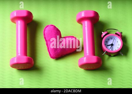 Amour du sport et de l'entraînement précoce. Décoration de coeur près des cloches en plastique sur le tapis de yoga. Concept d'entraînement matinal. Haltères de couleur rose à côté du réveil et coeur en peluche sur fond vert Banque D'Images