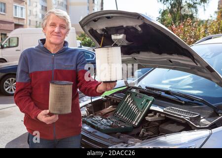 Pilote caucasien mature montrant des cartouches de filtre à air neuves et usagées pour moteur de voiture debout devant son véhicule avec capot ouvert Banque D'Images