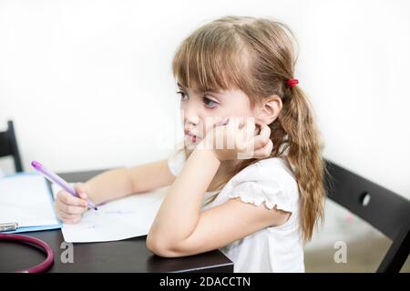 Petite fille caucasienne avec triste regard assis au bureau avec stylo à la main, leçon ennuyeuse, fond blanc Banque D'Images