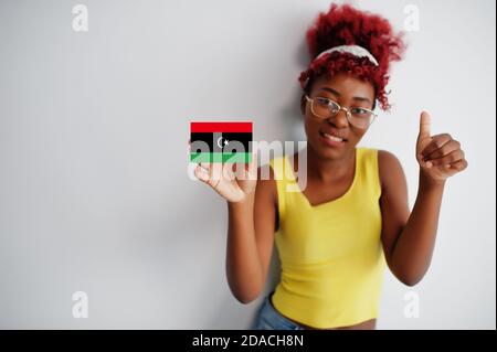 Femme africaine aux cheveux afro, porter un maillot jaune et des lunettes, tenir le drapeau libyen isolé sur fond blanc, montrer le pouce vers le haut. Banque D'Images