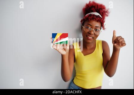 Femme africaine aux cheveux afro, porter un maillot jaune et des lunettes, tenir le drapeau des Seychelles isolé sur fond blanc, montrer le pouce vers le haut. Banque D'Images