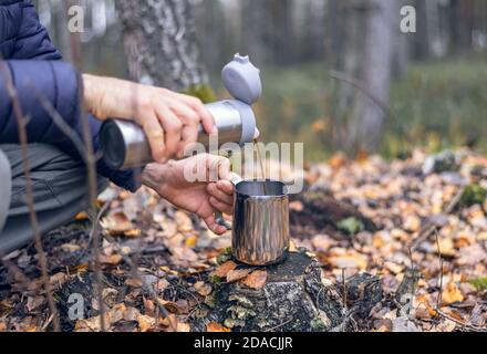 La personne (à la main uniquement) verse du café chaud d'un thermos dans une tasse en acier inoxydable sur une souche dans une forêt pleine de feuilles mortes le jour de l'automne aт. Banque D'Images