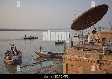 Varanasi, Inde, octobre 2011. Un homme assis dans un ghat sur le fleuve Ganges avec des bateaux pleins de pèlerins en arrière-plan. Banque D'Images