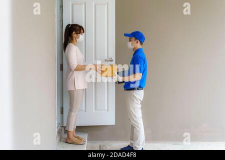 Homme de livraison asiatique portant un masque et un gant en uniforme bleu tenant une boîte en carton devant la maison et une femme acceptant une livraison de boîtes de d Banque D'Images