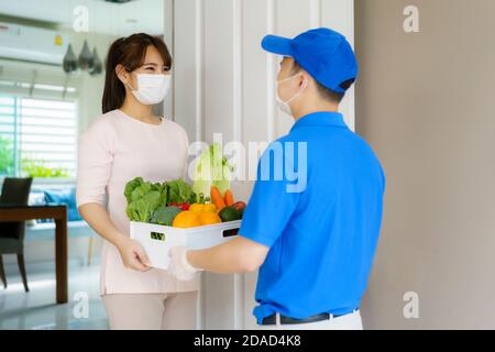 Une femme asiatique costumée portant un masque facial reçoit une boîte d'épicerie de nourriture, de fruits et de légumes de l'homme de livraison devant la maison pendant le temps de hom Banque D'Images