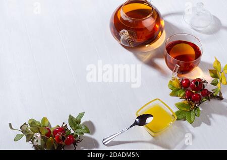 Thé d'automne confortable avec thé aux hanches roses dans une théière avec tasse, miel, branche de rose pour chien avec ombres sur une table en bois blanc clair et doux, espace de copie. Banque D'Images