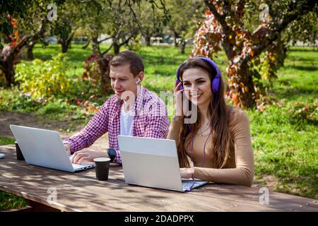 Un gars et une fille s'assoient à une table en bois dans le parc et travaillent sur des ordinateurs portables avec des écouteurs. Freelance travaille à l'extérieur. Travailler à l'extérieur du bureau en raison de Banque D'Images