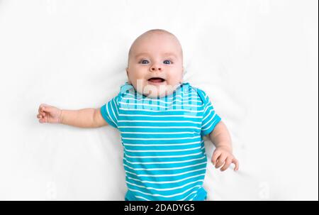 Un enfant positif portant un t-shirt rayé bleu repose sur un lit blanc, souriant et regardant l'appareil photo. Bonne enfance. Vue de dessus Banque D'Images