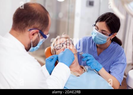 Stomatolog avec assistant de pose de joint dentaire sur les dents de femme âgée. Patient âgé pendant un examen médical avec un dentiste dans un cabinet dentaire équipé d'un équipement orange. Banque D'Images