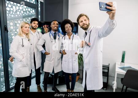 Une équipe souriante de cinq médecins multiethniques, africains, caucasiens et indiens, debout au centre médical moderne prenant un selfie. Groupe de Banque D'Images