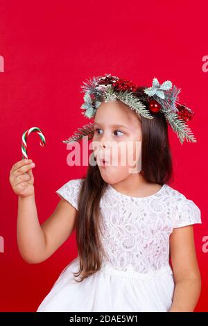 Gros plan d'une fille surprise dans une couronne de Noël regardant une canne au caramel dans sa main, sur un fond rouge Banque D'Images