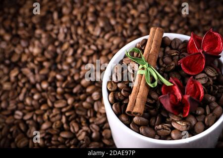 Tasse blanche pleine de grains de café sur fond de grains de café avec des fleurs rouges et de la cannelle. Espresso du matin. Tasse à café. Banque D'Images