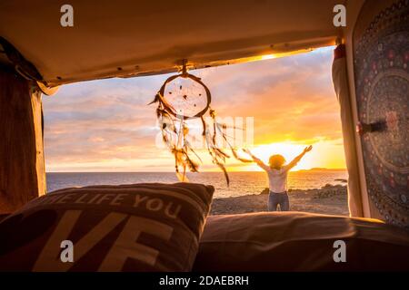 Concept de voyage et de liberté avec une femme heureuse debout à l'extérieur minibus admirez le coucher du soleil sur la plage - dreamcatcher en premier plan et soleil en arrière-plan Banque D'Images