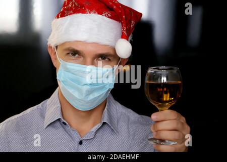Homme dans le masque médical et chapeau du Père Noël avec un verre de vin en main. Célébration de Noël pendant la pandémie du coronavirus Banque D'Images