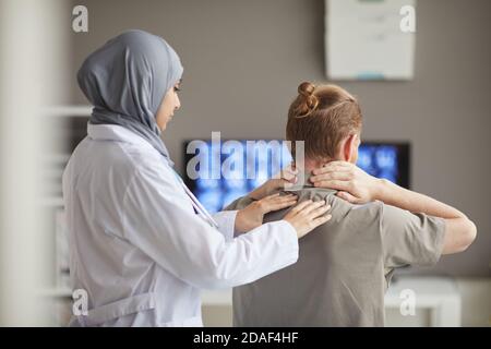 Vue arrière de la femme médecin en hijab examinant le cou du patient pendant qu'il est assis à son siège hôpital Banque D'Images