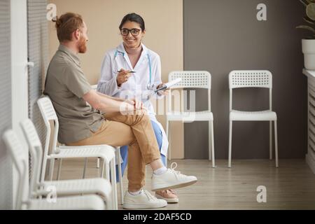 Jeune infirmière en lunettes souriant et parlant au patient pendant ils sont assis sur des chaises dans le couloir de l'hôpital Banque D'Images