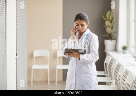 Jeune femme médecin en manteau blanc parlant sur téléphone portable et d'utiliser une tablette numérique pour travailler en ligne sur le corrior à l'hôpital Banque D'Images