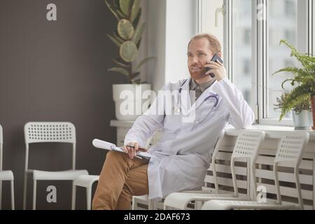 Jeune homme docteur en manteau blanc assis sur la chaise à le couloir de l'hôpital et de parler sur le téléphone mobile Banque D'Images