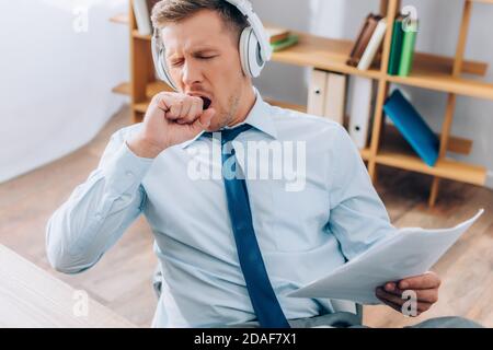 Un jeune homme d'affaires dans un casque bâillant tout en tenant des documents au bureau Banque D'Images