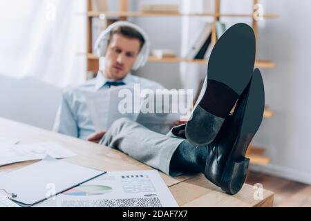Homme d'affaires avec des jambes sur la table, utilisant des écouteurs et travaillant avec documents sur un arrière-plan flou au bureau Banque D'Images