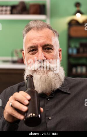 Un homme caucasien plus âgé à barbe, modèle barbu, dans un bar rétro hipster, boit une bouteille de bière, photo portrait. Banque D'Images