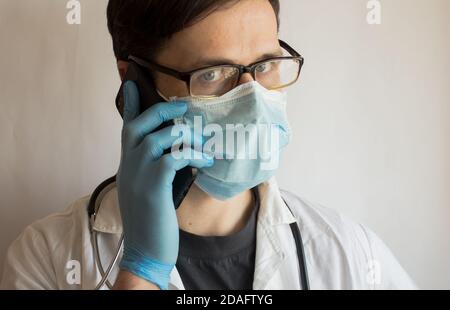 Un jeune beau médecin portant des lunettes et un masque médical donne une consultation au téléphone. Banque D'Images