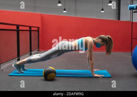 Ajuster la femme caucasienne portant un masque de visage exécutant l'exercice de planche dans la salle de gym Banque D'Images
