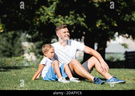 Père souriant assis près de son fils dans des vêtements de sport tout en regardant loin dans le parc avec arbres flous sur fond Banque D'Images