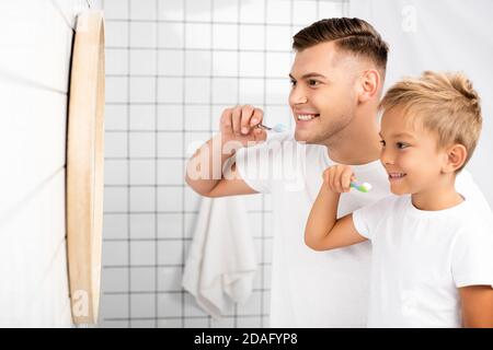 Un père et un fils souriants tenant des brosses à dents et regardant le miroir dans la salle de bains Banque D'Images