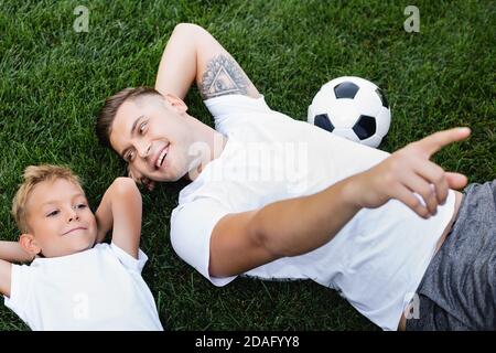 Vue en hauteur d'un père heureux pointant avec le doigt, tout en regardant un fils souriant allongé avec les mains derrière la tête sur l'herbe