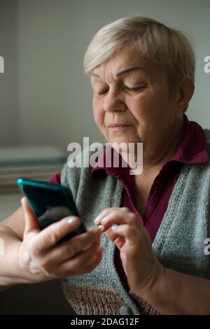 Une vieille femme utilise un téléphone cellulaire. Une femme âgée appelle sur un smartphone tout en étant assise dans une pièce à la maison. Banque D'Images