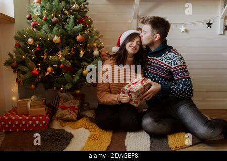 Elégant couple heureux échanger des cadeaux de noël sous l'arbre de noël avec des cadeaux et des lumières dans une salle décorée de fête. Une jeune famille s'embrasse et tient Banque D'Images