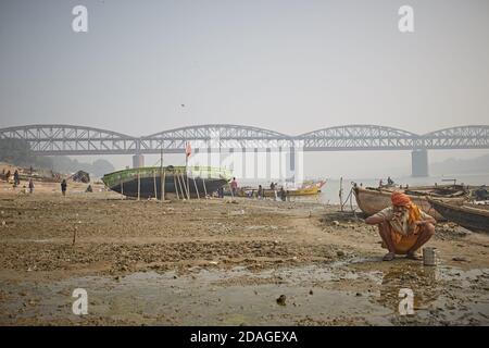 Varanasi, Inde, janvier 2016. Un homme brosse ses dents sur une plage de Ghat sur le fleuve Ganges avec un grand pont en métal en arrière-plan. Banque D'Images