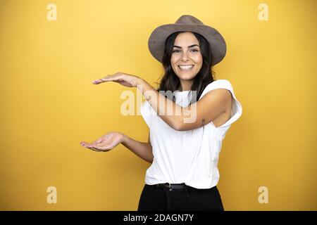 Belle femme portant un t-shirt blanc décontracté et un chapeau debout sur fond jaune gestante avec les mains montrant grand et grand signe de taille, mesure s Banque D'Images