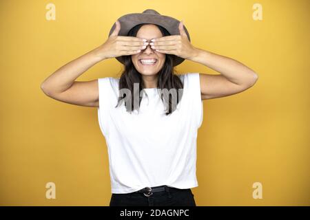 Belle femme portant un t-shirt blanc décontracté et un chapeau debout sur fond jaune couvrant les yeux avec les mains souriantes et drôles. Conc. Aveugle Banque D'Images
