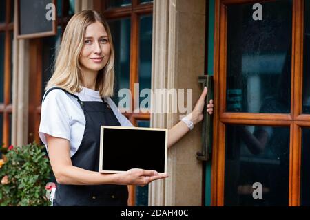 Jeune femme caucasienne de 30 ans avec cheveux blonds courts tenant une pancarte avec espace de copie près des portes du café. Rouvrir le restaurant après le coronavirus Banque D'Images